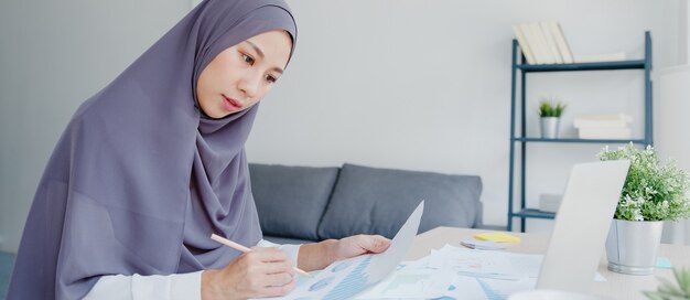Schöne asiatische muslimische Dame in Kopftuch-Freizeitkleidung mit Laptop im Wohnzimmer im Haus.