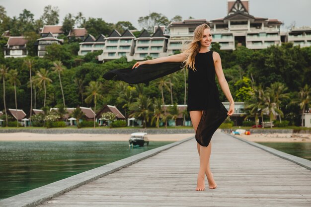 Schöne attraktive Frau gekleidet im schwarzen Kleid, das auf Pier im Luxusresorthotel, Sommerferien, tropischer Strand aufwirft