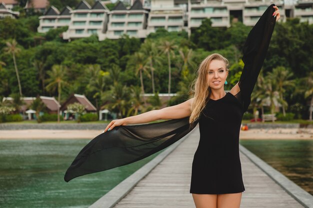 Schöne attraktive Frau gekleidet im schwarzen Kleid, das auf Pier im Luxusresorthotel, Sommerferien, tropischer Strand aufwirft