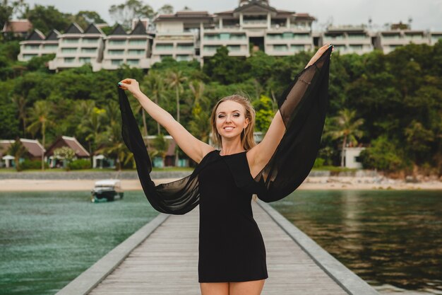 Schöne attraktive Frau gekleidet im schwarzen Kleid, das auf Pier im Luxusresorthotel, Sommerferien, tropischer Strand aufwirft