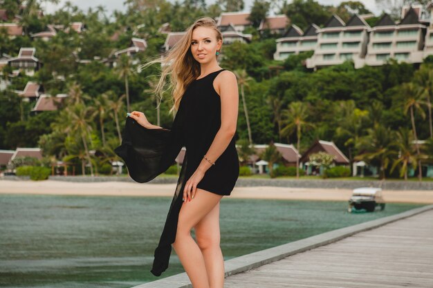 Schöne attraktive Frau gekleidet im schwarzen Kleid, das auf Pier im Luxusresorthotel, Sommerferien, tropischer Strand aufwirft