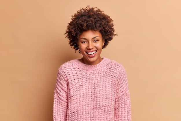 Schöne aufrichtige Frau mit Afro-Haar lächelt breit hat weiße perfekte Zähne verbirgt keine positiven Emotionen trägt Strickpullover isoliert über brauner Wand