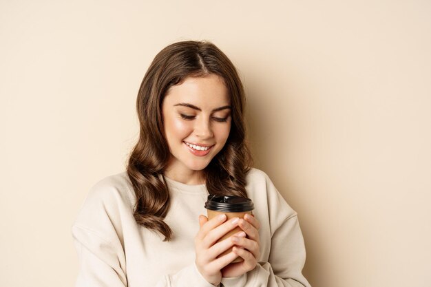 Schöne authentische Frau, die lächelt, eine warme Tasse Kaffee in der Hand hält und glücklich aussieht, über beigem Hintergrund steht.