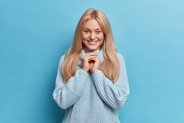 Schöne blonde europäische Frau lächelt sanft, hält die Hände zusammen, während Grübchen auf den Wangen einen gestrickten Pullover tragen, der etwas Angenehmes hört