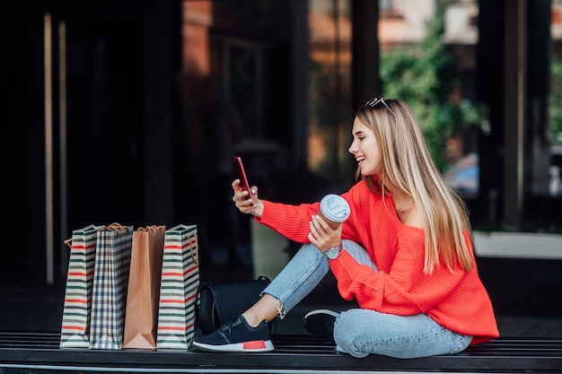 Kostenloses Foto schöne blonde frau, die auf der straße sitzt und von einkaufstüten umgeben ist und am telefon schaut.