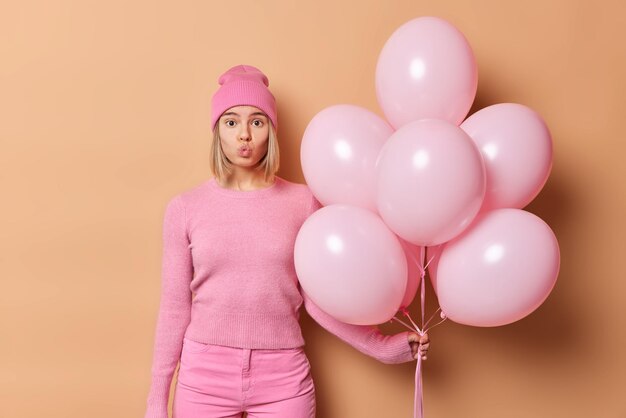 Schöne blonde Frau hält die Lippen rouded sendet Luftkuss an die Kamera kommt auf Geburtstagsfeier hält Haufen aufgeblasener Heliumballons trägt rosa Kleidung isoliert über braunem Hintergrund Feierkonzept