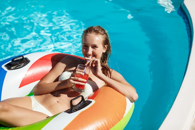 Schöne blonde Frau lächelnd, Cocktail trinkend, im Pool schwimmend