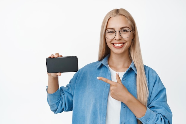 Schöne blonde frau mit brille, die eine app demonstriert, die mit dem finger auf das handy zeigt und einen weißen hintergrund lächelt