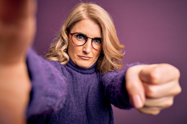 Kostenloses Foto schöne blonde frau mittleren alters mit pullover und brille macht ein selfie vor der kamera, genervt und frustriert, schreiend vor wut, verrückt und schreiend mit erhobener hand