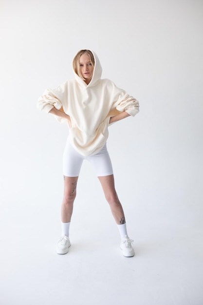 Schöne blonde Frau posiert in weißem Hoodie und Leggings gegen weißen Hintergrund