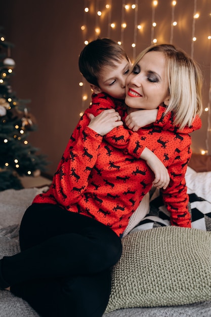 Schöne blonde Frau umarmen ihr zartes Sohnlied auf dem Bett vor einem Weihnachtsbaum