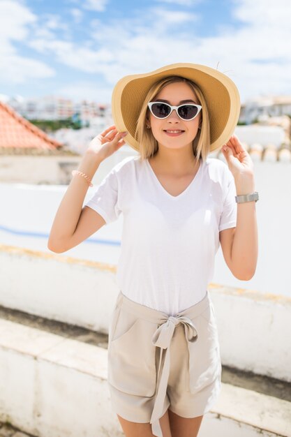 Schöne blonde junge Frau, die Hut und Sonnenbrille trägt, die auf der Straße gehen