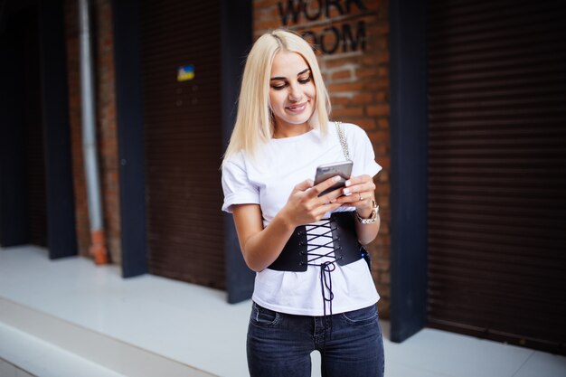 Schöne blonde kaukasische Frau mit Headset, das vor Ziegelmauer steht, die eine Textnachricht tippt und in ihrem Lieblingskaffee genießt.