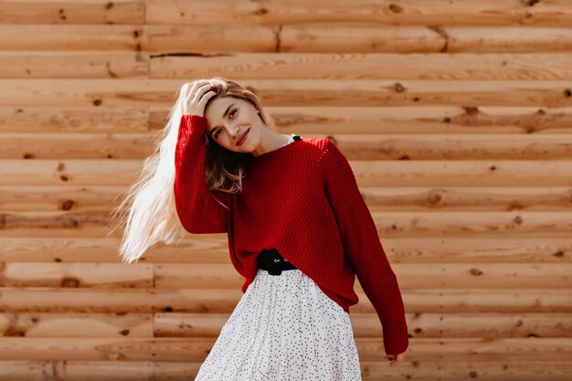 Schöne Blondine im trendigen roten Pullover mit viel Spaß draußen. Fröhliche junge Frau, die glücklich auf der Holzwand aufwirft.