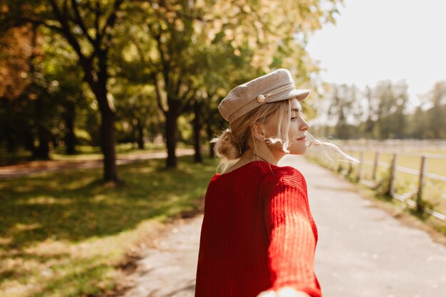 Schöne Blondine in trendigen Kleidern, die spielerisch darum bittet, ihr im Park zu folgen. Hübsches Mädchen, das sonniges Wetter im Freien genießt.