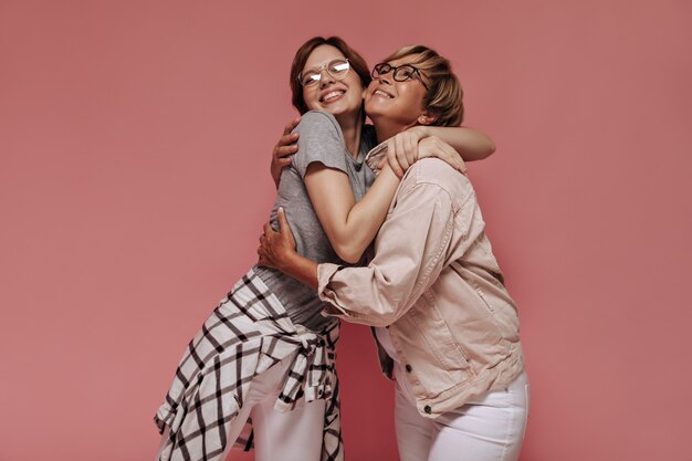 Schöne brünette Dame mit Brille im grauen T-Shirt, weißen Hosen und kariertem Hemd, das lächelnde blonde Frau in beige Jacke auf rosa Hintergrund umarmt.