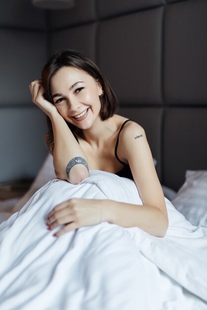 Schöne brünette Frau des langen Haares auf weißem Bett im weichen Morgenlicht unter der Bettdecke