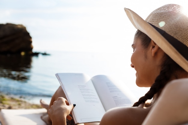 Schöne brünette Frau im Hutlesebuch, am Strand liegend