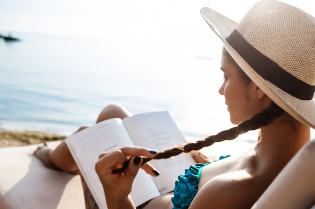 Schöne brünette Frau im Hutlesebuch, am Strand liegend