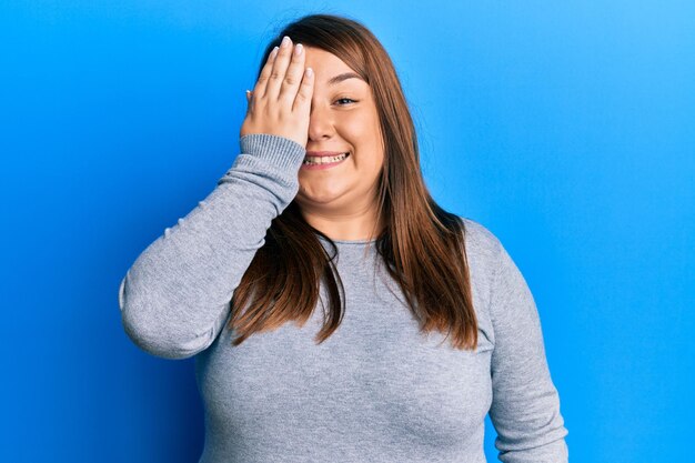 Schöne brünette Frau in Übergröße in lässiger Kleidung, die ein Auge mit der Hand bedeckt, selbstbewusstes Lächeln im Gesicht und überraschende Emotionen.