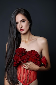 Schöne brünette frau mit einem strauß roter rosenblüten im roten korsett. langes haar, nacktes, schlankes körperkunstporträt