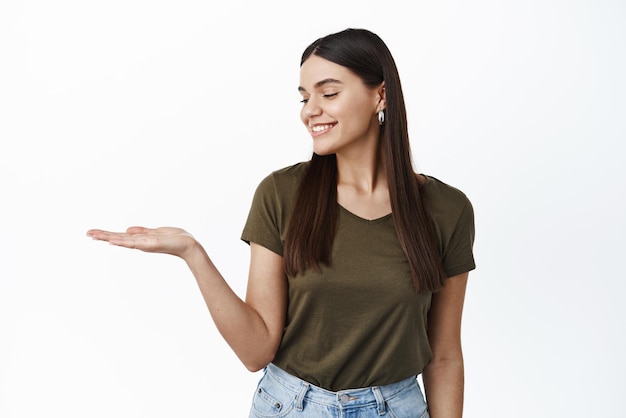 Schöne brünette Frau mit natürlichem Make-up in grünem T-Shirt, das Ihr Logo-Produkt auf der Handfläche hält und leere Hand auf weißem Hintergrund des Kopierraums betrachtet