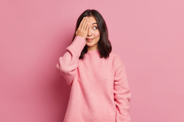 Schöne brünette junge asiatische Frau mit östlichem Aussehen bedeckt Augen mit Hand versteckt Gesicht lächelt angenehm trägt lässig gestrickte Strickposenposen