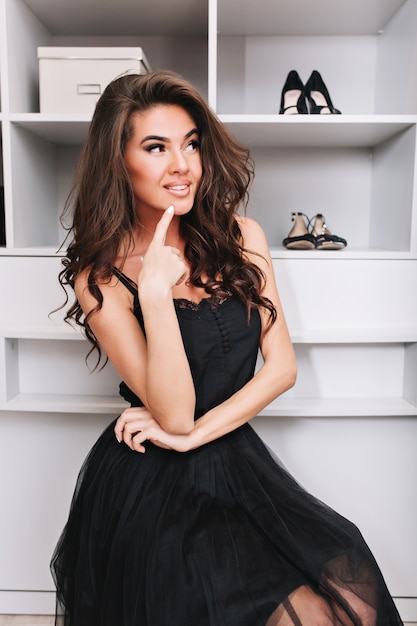Schöne brünette junge Frau, die nachdenklich in der Umkleidekabine sitzt und über die Wahl der Kleidung nachdenkt. Sie ist glücklich und trägt ein schwarzes Outfit.