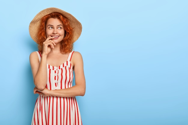 Schöne charmante junge Dame mit Ingwerhaar, das im Sommerkleid und im Strohhut aufwirft