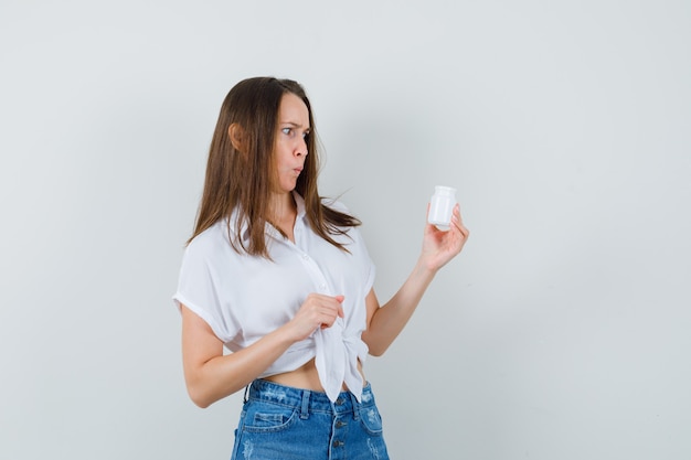 Schöne Dame, die Flasche der Pillen in der weißen Bluse, in den Jeans betrachtet und verwirrt schaut. Vorderansicht.