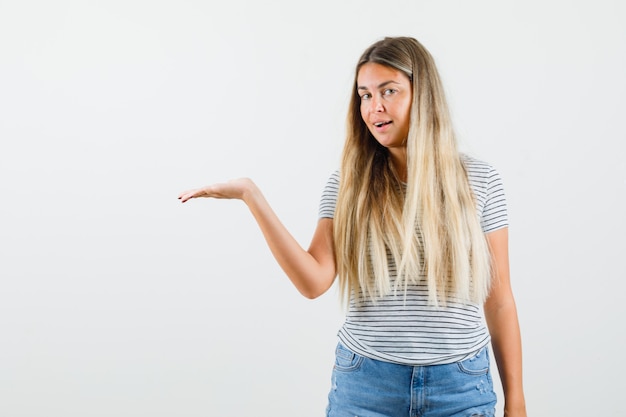 Schöne Dame im T-Shirt, die ihre offene Handfläche hebt, um etwas beiseite zu zeigen und zufrieden zu sehen, Vorderansicht.