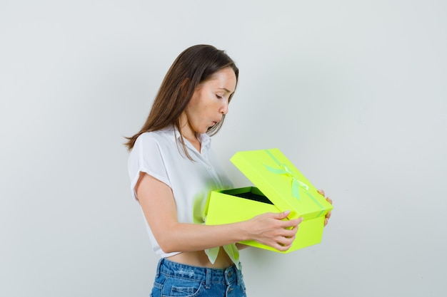 Schöne Dame in der weißen Bluse, die gelbe Geschenkbox öffnet und ungeduldig schaut, Vorderansicht.