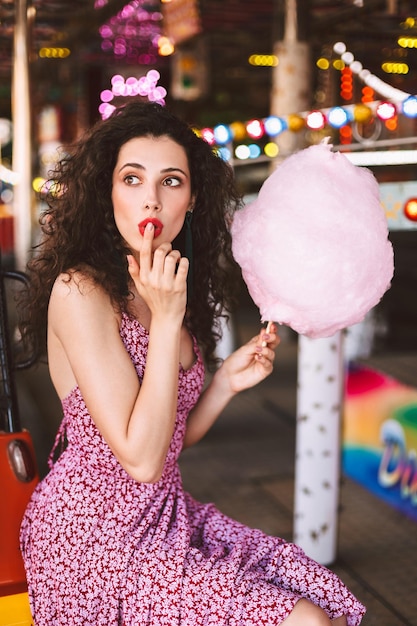 Schöne dame mit dunklem, lockigem haar im kleid, die mit zuckerwatte in der hand sitzt und verträumt zur seite schaut, während sie zeit im vergnügungspark verbringt