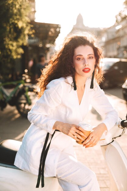 Schöne Dame mit dunklem lockigem Haar in weißem Kostüm sitzt auf weißem Moped mit einer Tasse Kaffee zum Mitnehmen und blickt nachdenklich in die Kamera auf der Straße