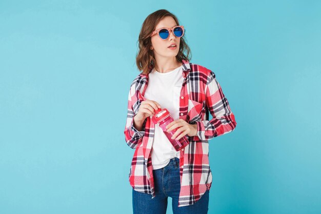 Schöne Dame mit Sonnenbrille, die mit rosa Sportflasche auf blauem Hintergrund steht Foto eines netten Hipster-Mädchens in kariertem Hemd und Jeans