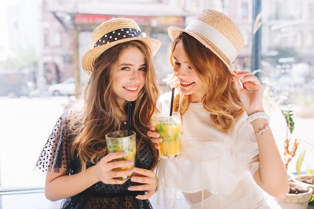 Schöne Damen tragen ähnliche Strohhüte, die gemeinsam Spaß haben und am Sommertag eisige Fruchtcocktails genießen