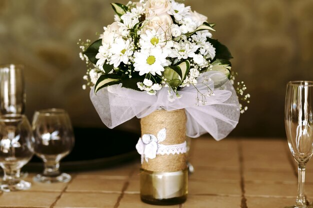 Schöne dekorative Vase mit üppigem Bouquet von Gänseblümchen und rosa Rosen und Grün auf dem Tisch unter den Gläsern Champagner und Schnaps.