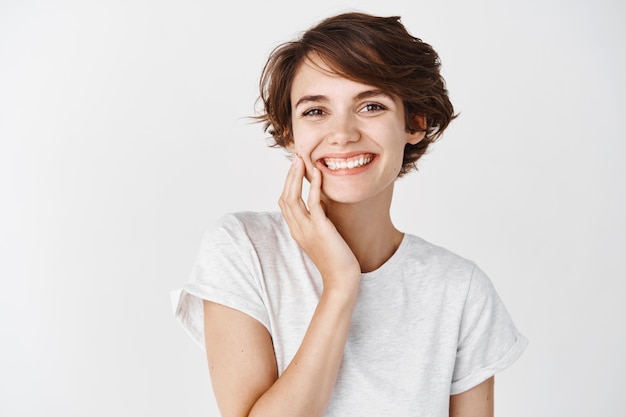 Schöne ehrliche Frau mit kurzen Haaren und ohne Make-up, die saubere Gesichtshaut berührt und lächelt, im T-Shirt auf weißer Wand stehend