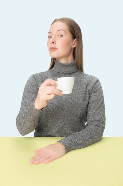 Schöne einsame Frau, die am blauen Studio sitzt und traurig schaut, die Tasse Kaffee in der Hand haltend. Nahaufnahme getöntes Porträt im Minimalismusstil