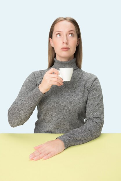 Schöne einsame Frau, die im blauen Studio sitzt und traurig schaut, die Tasse Kaffee in der Hand haltend.