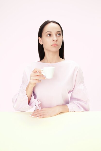 Schöne einsame Frau, die im rosa Studio sitzt und traurig schaut, die Tasse Kaffee in der Hand haltend.