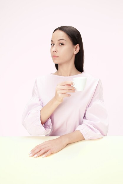 Schöne einsame Frau, die im rosa Studio sitzt und traurig schaut, die Tasse Kaffee in der Hand haltend.