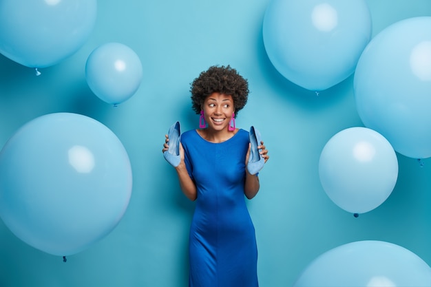 Schöne elegante Frau im blauen Kleid, hält Schuhe mit hohen Absätzen, genießt das Feiern, hat Spaß auf der Party, schaut mit einem Lächeln beiseite, posiert um Luftballons. Die afroamerikanische Dame trägt ein modisches Outfit