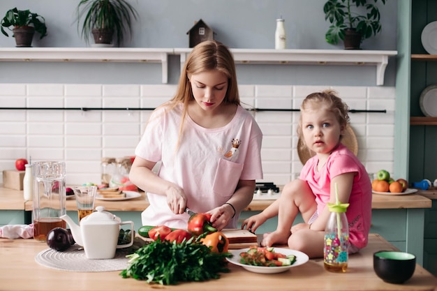 Schöne Eltern kochen Abendessen in der Küche mit ihrem kleinen süßen Kind Tochter sitzt auf dem Tisch und isst, während die Mutter Pfeffer schneidet Glückliche Frau, die zusammen mit dem Kind Mittagessen aus Gemüse zubereitet