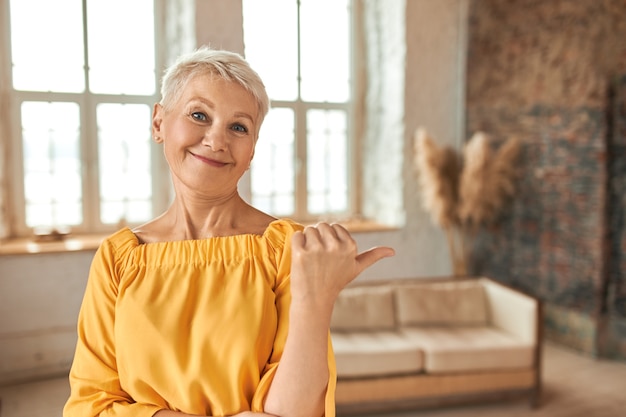 Schöne erfolgreiche immobilienmaklerin mittleren alters mit pixie-haarschnitt, die daumen hoch geste macht, finger auf gemütliches wohnzimmer mit stilvollem innendesign zeigt und wohnung zum verkauf anbietet