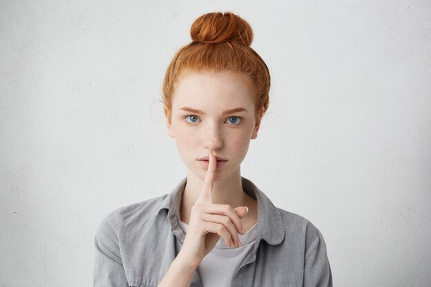 Schöne ernsthafte junge Frau mit Haarknoten gekleidet, die kausal Finger an ihren Lippen hält und bittet, keinen Lärm zu machen oder Zunge zu halten und einige private vertrauliche Informationen zu verbergen. Streng geheim