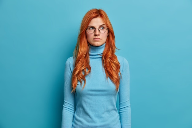 Kostenloses Foto schöne ernsthafte rothaarige frau konzentriert mit nachdenklichem ausdruck beiseite trägt runde brille und rollkragenpullover.