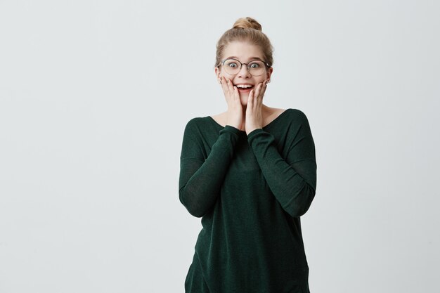 Schöne erstaunte Frau mit blondem Haarknoten und Brille kann ihren Augen nicht trauen, froh, unerwartetes Geschenk von Freund zu erhalten, hat glücklichen und überraschten Ausdruck. Glückskonzept.