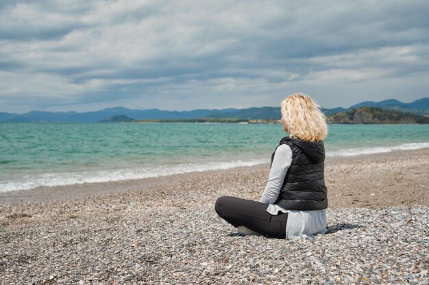 Schöne erwachsene blonde Frau in Sportbekleidung sitzt an einem Kiesstrand in der Nähe des azurblauen Ägäischen Meeres natürliches Sonnenlicht mittags Blick von hinten vor dem Hintergrund von Meer und Wellen Rückansicht