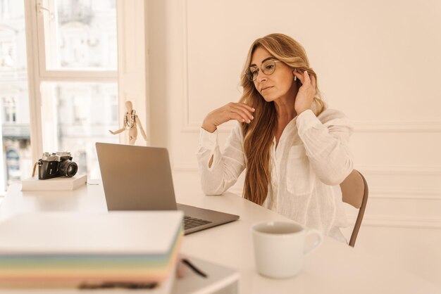 Schöne erwachsene kaukasische Geschäftsfrau, die am Computer arbeitet oder studiert, während sie am Tisch im hellen Büro sitzt Freiberufliches Konzept